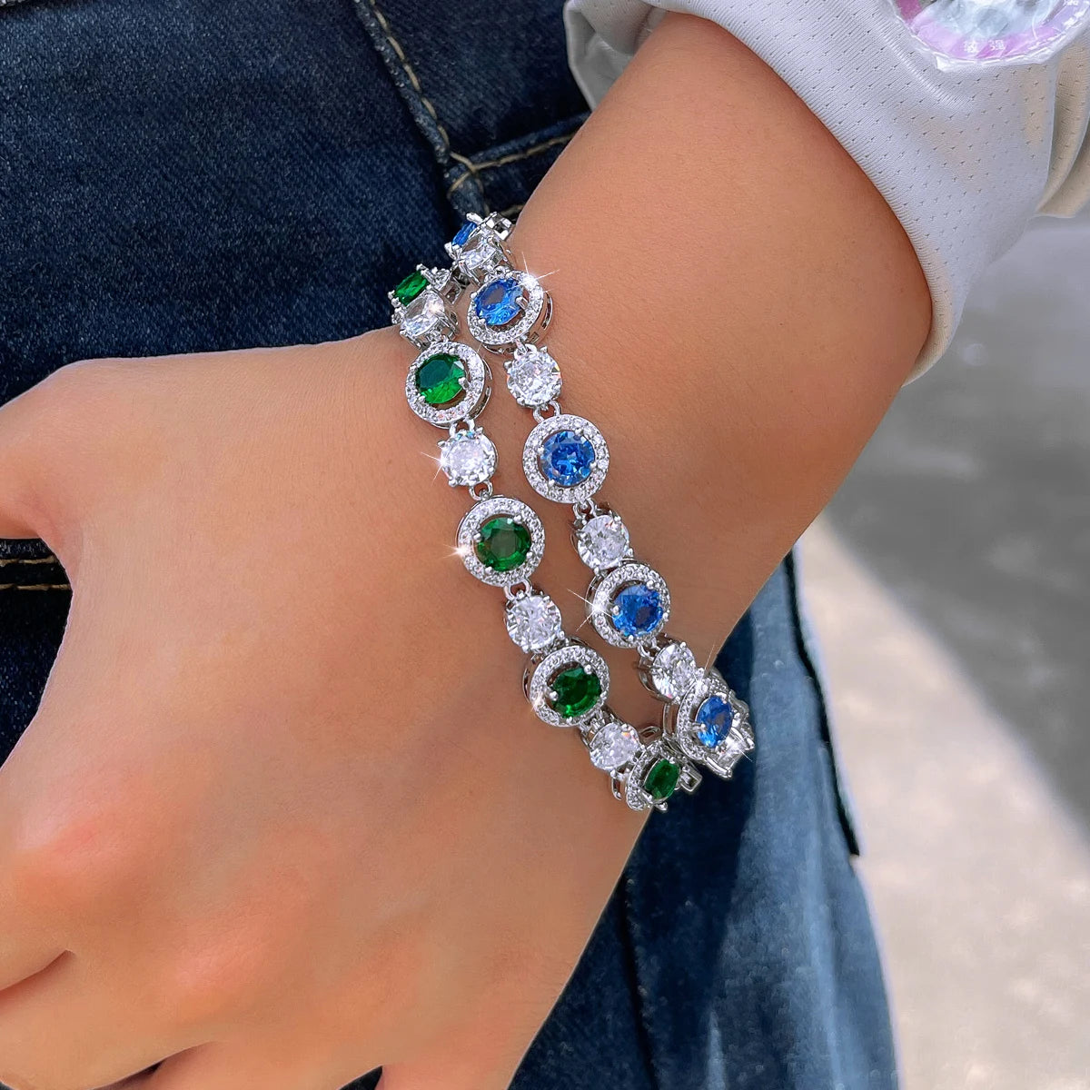 Silver-tone tennis bracelets with round blue, green, and clear stones worn on a hand