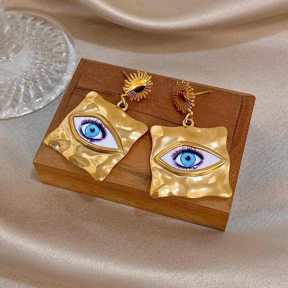 Gold-tone evil-eye drop earrings on wooden tray: blue eye enamel in hammered squares