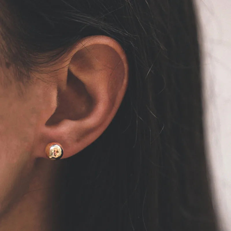 Small polished gold-tone ball stud earring worn on an earlobe