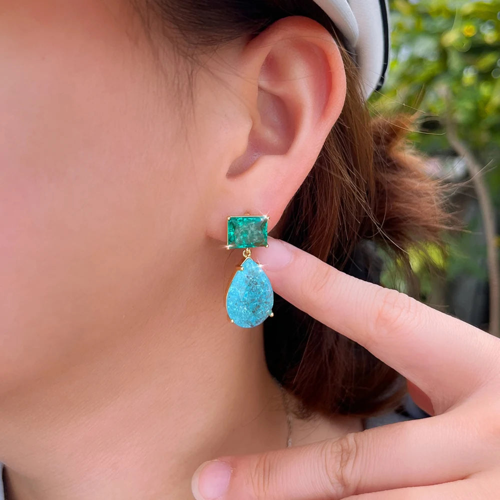 Green square and blue teardrop earring worn beside a finger for scale
