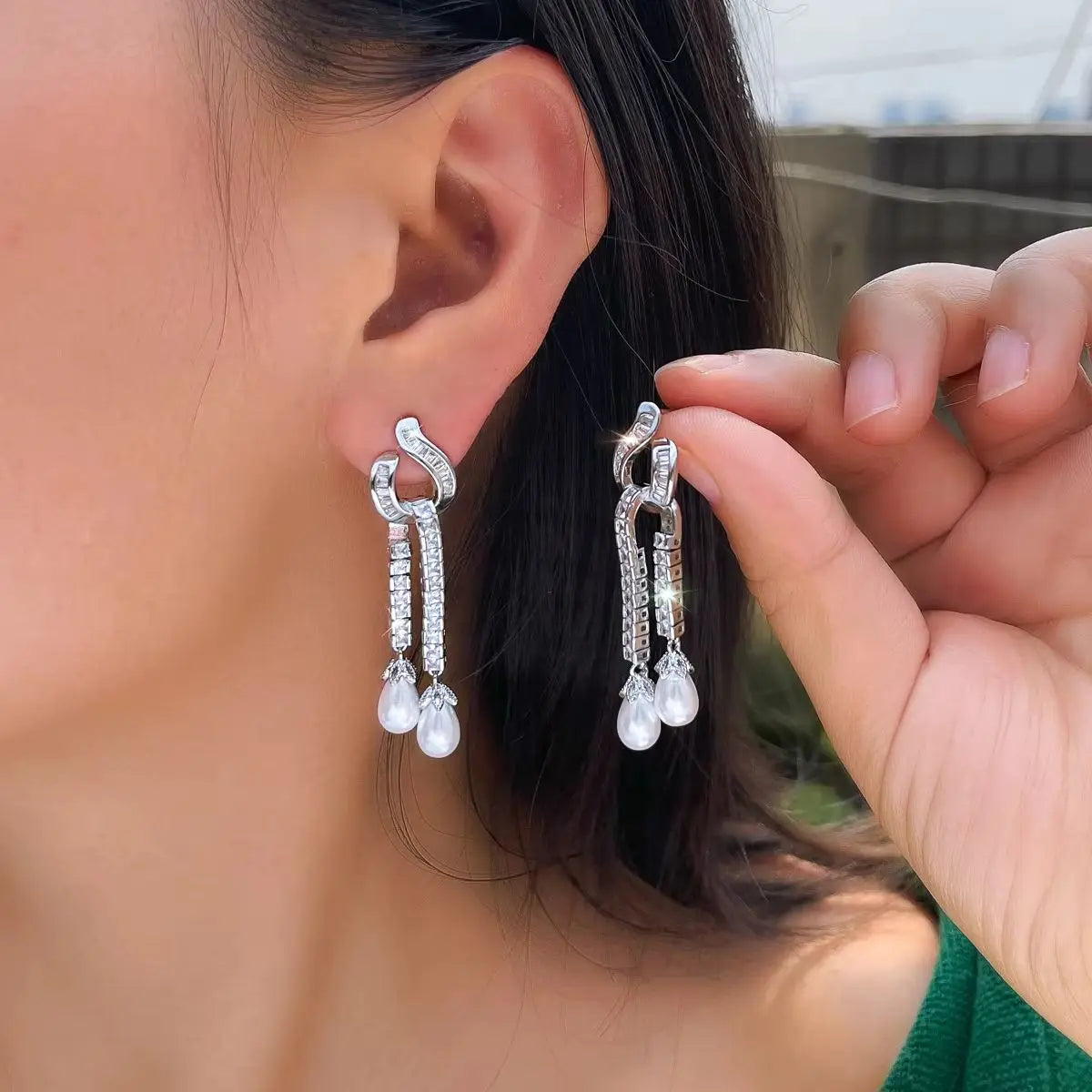 Silver-tone zirconia dangle earrings with two pearl drops, shown worn and held beside the ear.