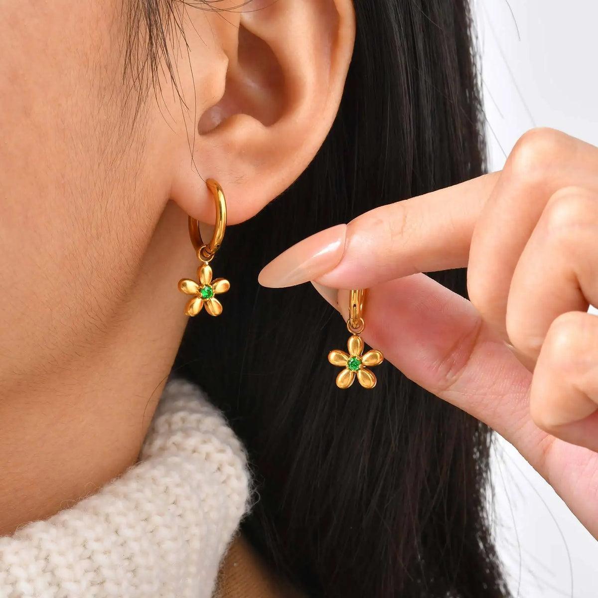 Gold flower charm hoop earrings worn and held up, with green stone centers.