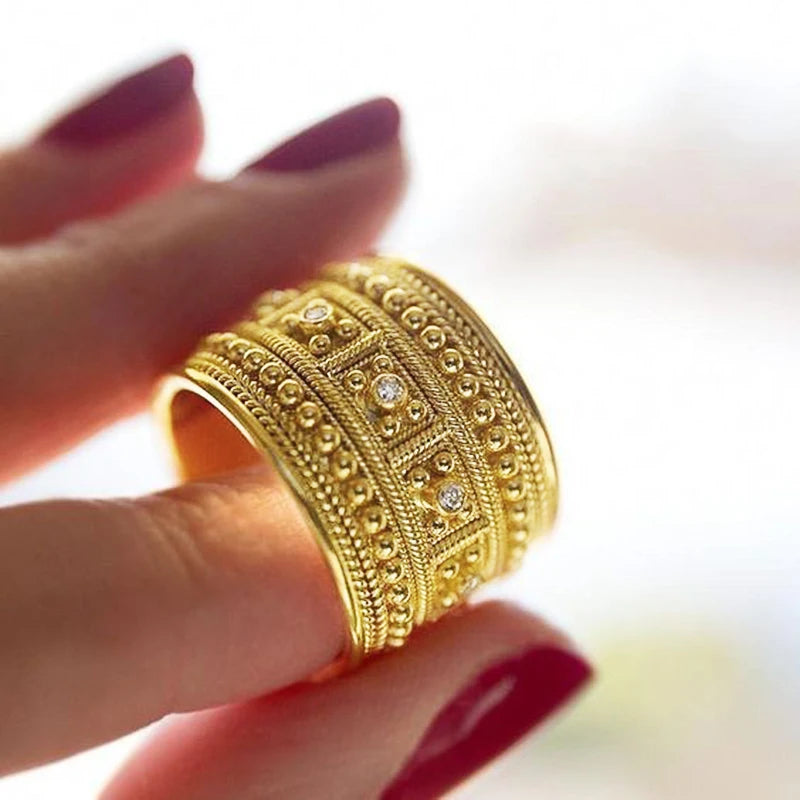 Gold-tone wide band ring worn on a finger, with detailed beading and clear stones