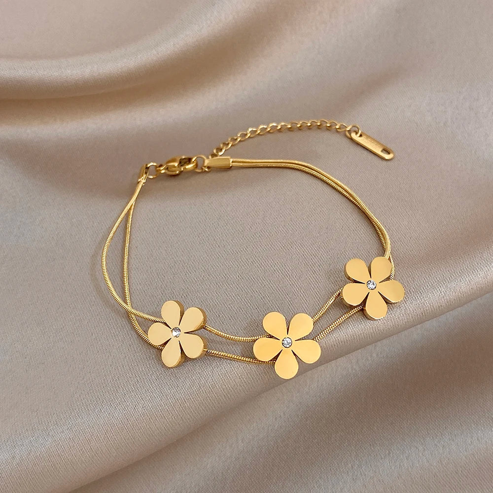 Gold double-layer snake chain bracelet with five flower charms and crystal centers, laid on fabric